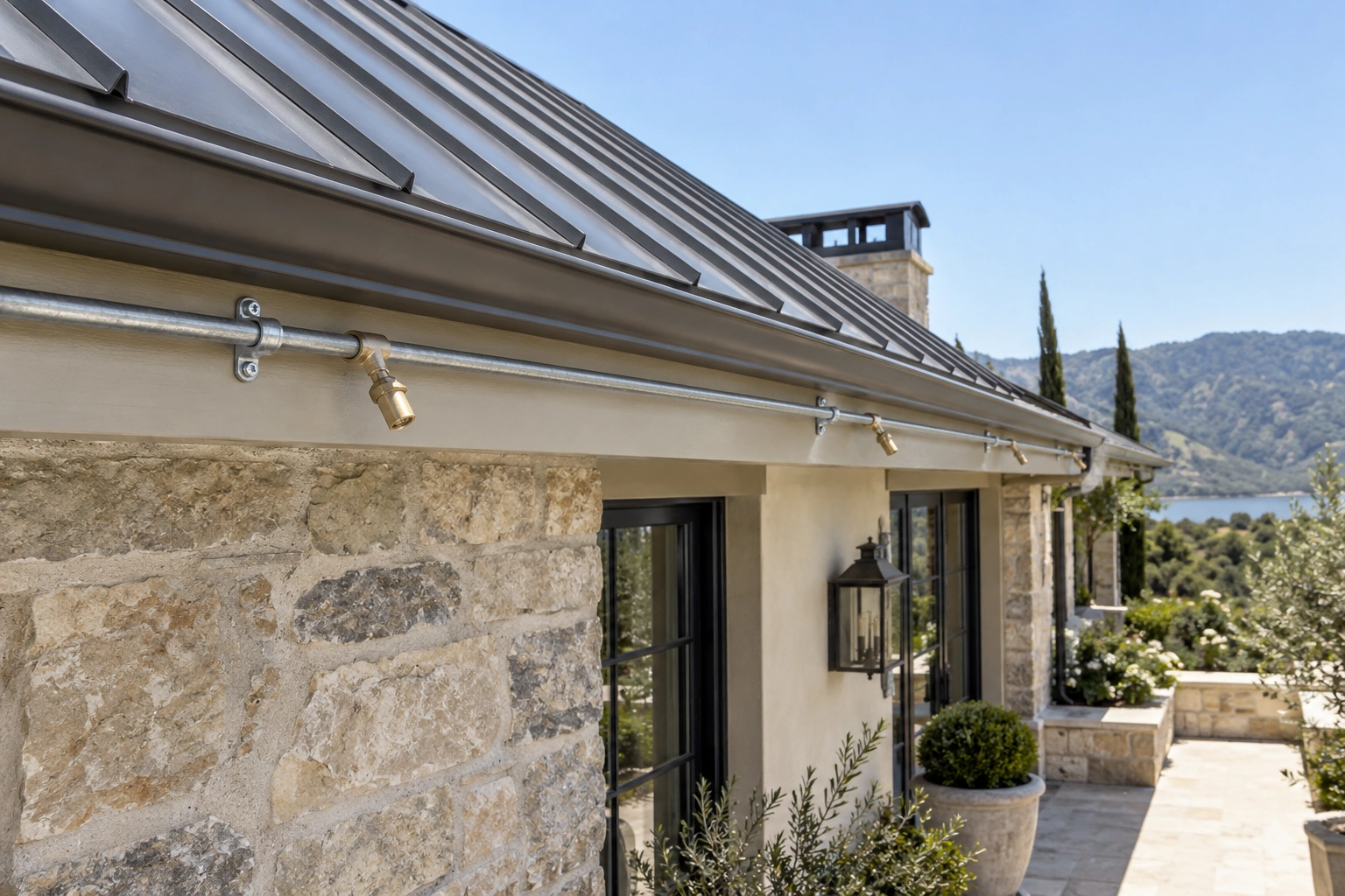 Supported metal pipe with connected brass exterior spray nozzles running along a luxury residence’s fascia